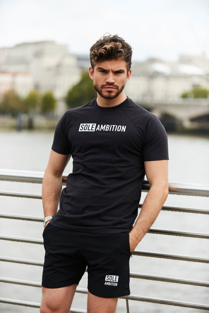 Man wearing matching set of black exercise t-shirt and shorts from Sole Ambition, posing on bridge, river in the background.