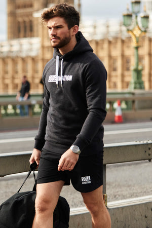 Man wears men's black gym shorts and fitted hoodie set from Sole Ambition walking to gym with houses of parliament in background
