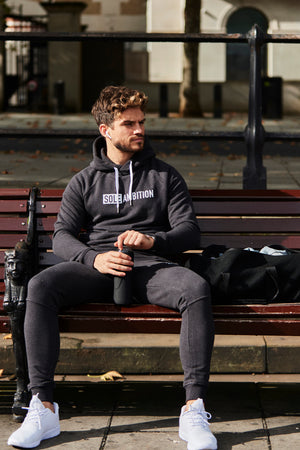 Man wearing a sole ambition fitted black cotton joggers and hoodie listens to music on a bench in London